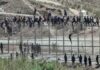 People climbing fence watched by security personnel below