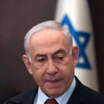 Man in suit with Israeli flag in background.