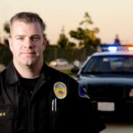 A police officer in uniform stands in front of a police car with lights on