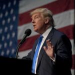 A man in a suit speaking at a podium with an American flag in the background