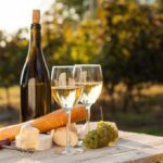A bottle of white wine, two glasses, cheese, and grapes on a wooden table in a vineyard