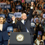 Walz Pulls Race Card In Pathetic Fraud Defense Two people celebrate on stage with supporters behind.