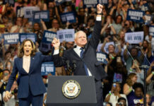 HOW Walz Programs Were DESIGNED for Theft Two people celebrate on stage with supporters behind.