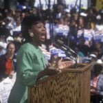 A woman passionately speaking at a political rally in front of a large crowd