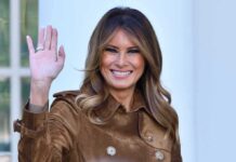 Melania Trump waving and smiling while wearing a stylish coat