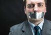 Man in business attire with duct tape over his mouth, conveying a message about silence