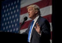 A man in a suit speaking at a podium with an American flag in the background