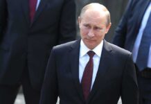 A serious-looking man in a suit walking outdoors with security personnel in the background