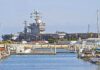 A large naval aircraft carrier docked in a harbor with smaller boats in the foreground