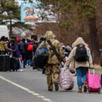 Group of people carrying luggage walking along a road with a soldier