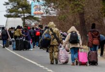 Group of people carrying luggage walking along a road with a soldier