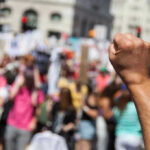 GLOBAL Protests ERUPT – UNSEEN RISKS Loom Raised fist in front of a crowd.