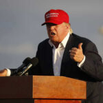 Trump TIGHTENS Housing Rules—WHO’S OUT? Person speaking at a podium wearing a red hat.