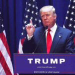 Media Hype Twists: New Global Poll Fuels Anti-Trump Narrative Man speaking at podium with USA flags behind him.