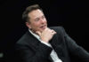 Times Square Balloon Protest Targets Musk’s AI Grok Man in suit smiling, resting chin on hand.