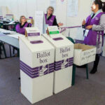 Few Cases Fuel NJ Election Debate Election polling station with ballot boxes and officials.