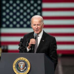 Man speaking at podium with US flag background.