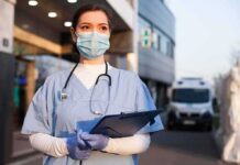 Healthcare worker in scrubs wearing mask and gloves holding a clipboard