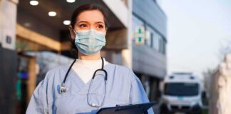 Healthcare worker in scrubs wearing mask and gloves holding a clipboard