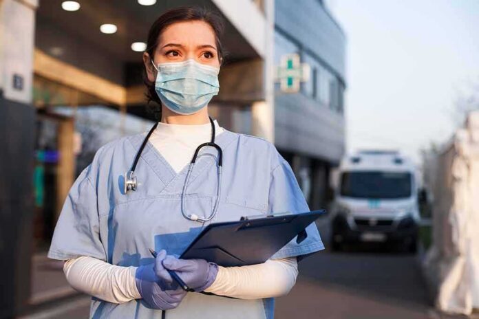 shutterstock_1714681855 Healthcare worker in scrubs wearing mask and gloves holding a clipboard