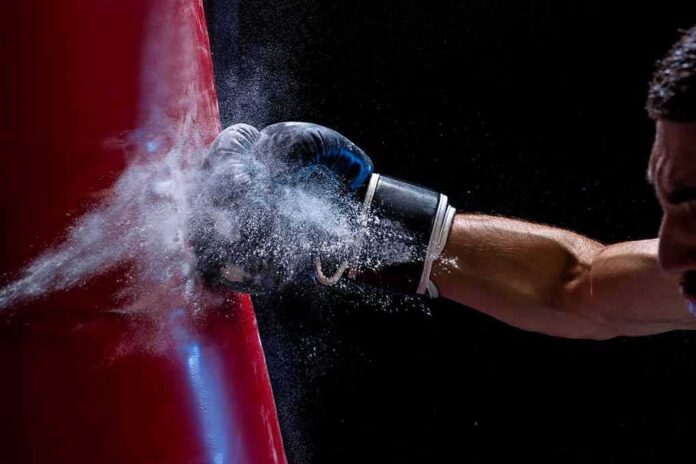 shutterstock_1135551392 Boxer’s gloved fist strikes a red punching bag, sending chalk dust into the air