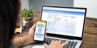Person using smartphone to approve bank transfer beside laptop with online banking