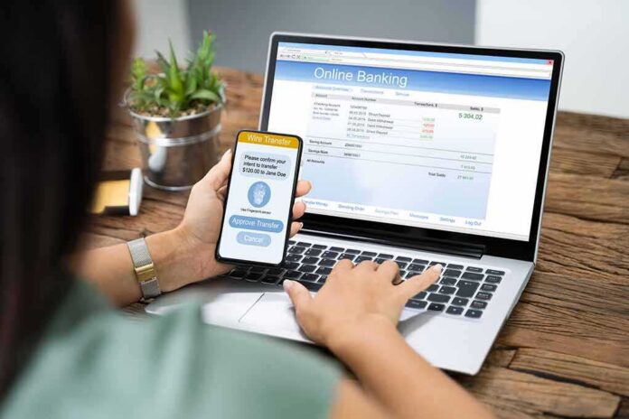 shutterstock_1542438851 Person using smartphone to approve bank transfer beside laptop with online banking
