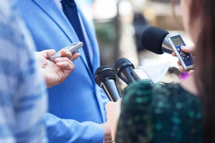 Reporters hold microphones toward a person in a blue suit