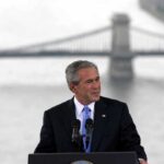 George W. Bush speaking at a podium with a bridge over water in the background