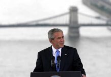 George W. Bush speaking at a podium with a bridge over water in the background