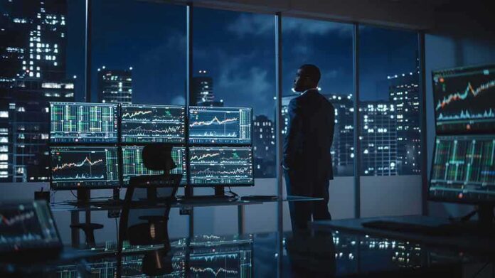 shutterstock_2088641509-1 Trader in a dark office with multiple stock monitors and city skyline at night